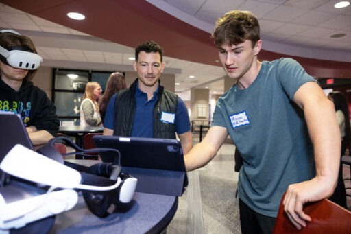 Two people discuss what is shown on the laptop in front of them while another person nearby is wearing a CR headset.