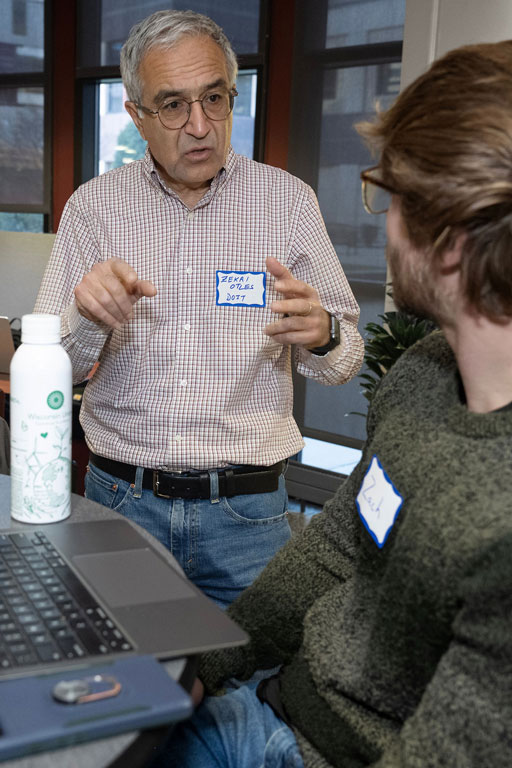 One man stands and talks with another man who is seated in front of a laptop.